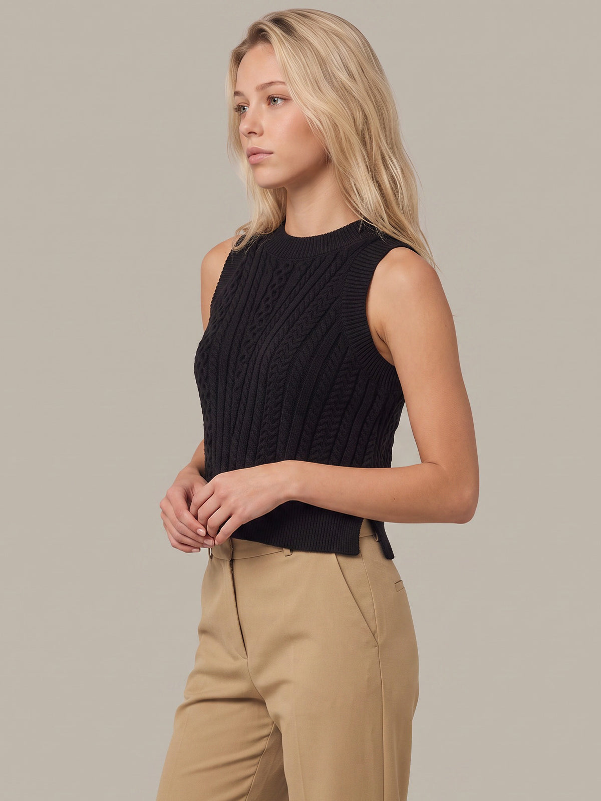 A blonde woman wearing a black sleeveless cable-knit mock neck top and beige trousers, photographed against a solid light gray studio background.