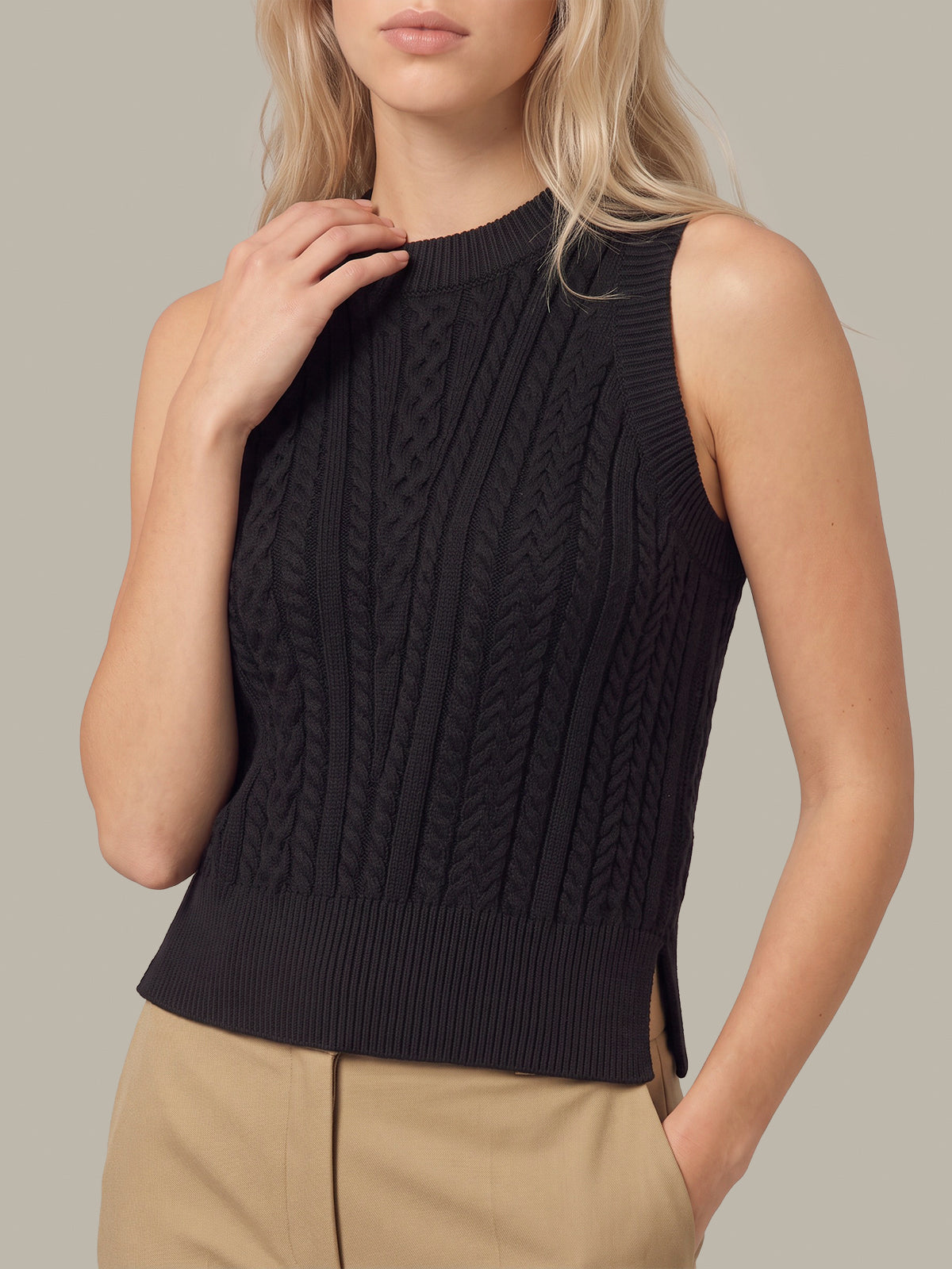 A woman wearing a sleeveless black cable-knit mock neck sweater vest paired with beige trousers, shown against a solid neutral background.