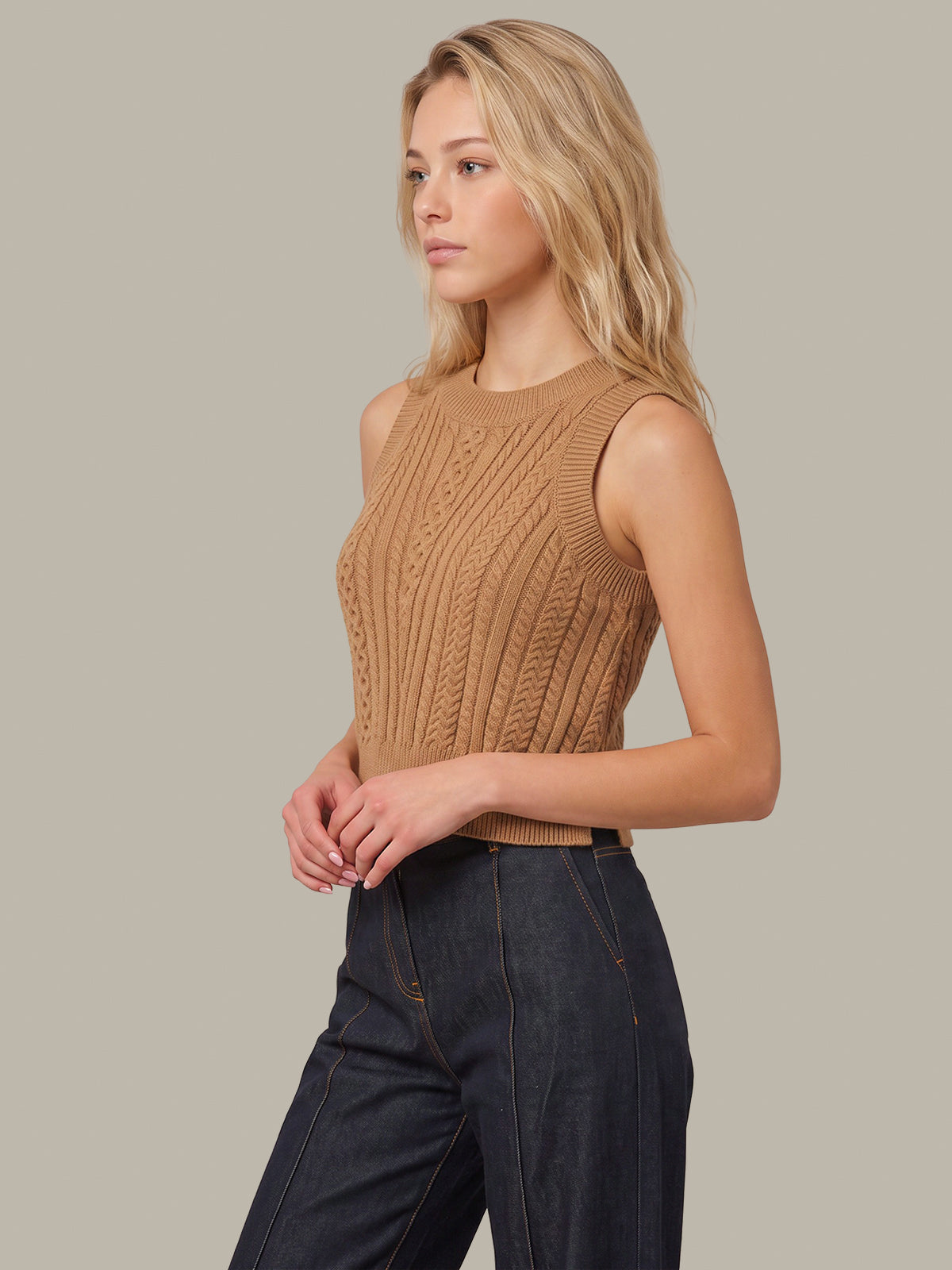 A woman wearing a sleeveless, ribbed knit sweater in camel brown, paired with dark blue jeans, photographed against a solid light gray background.