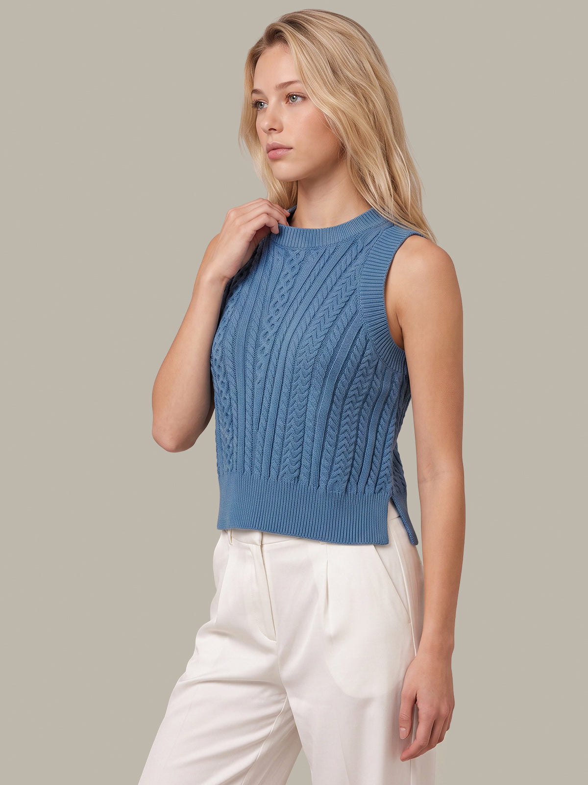 A blonde woman wearing a sleeveless blue cable-knit sweater vest and white trousers, posing against a solid light gray background.
