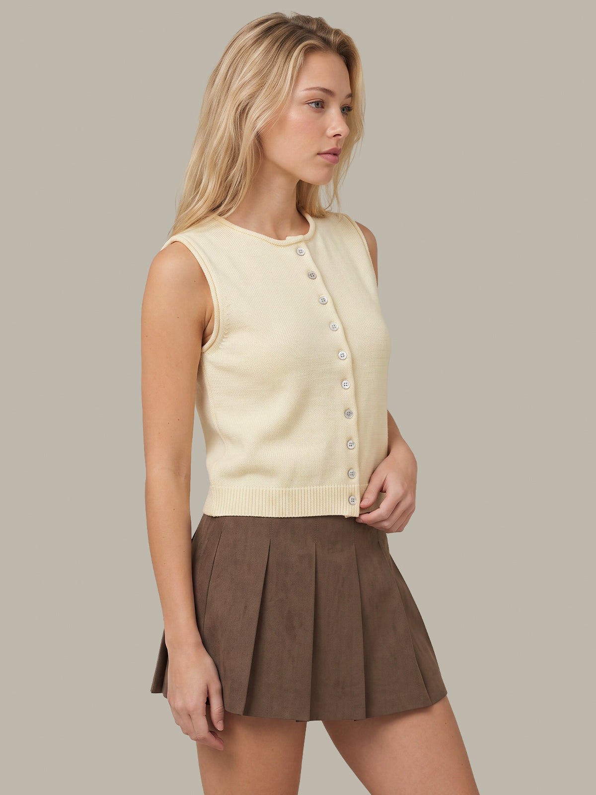 A woman modeling a cream sleeveless button-front knit top paired with a brown pleated mini skirt, set against a solid light gray studio background.