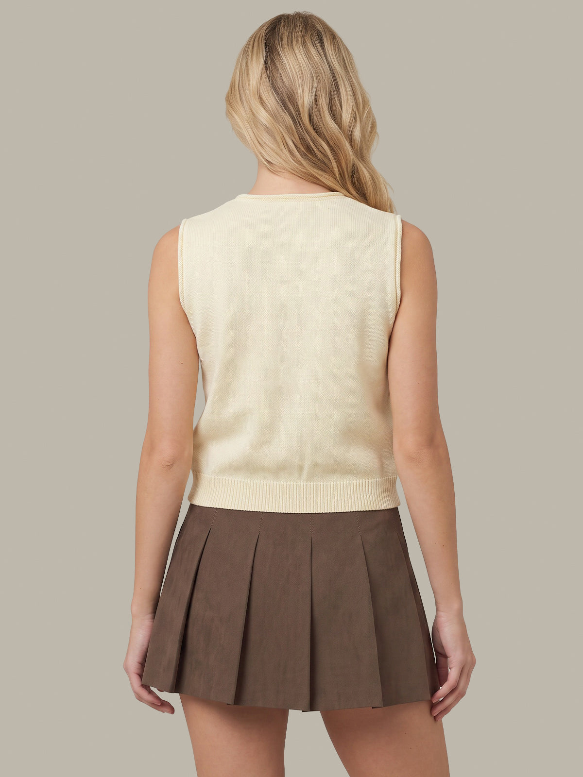 A woman shown from the back wearing a cream sleeveless knit top and a brown pleated mini skirt, against a solid taupe background.