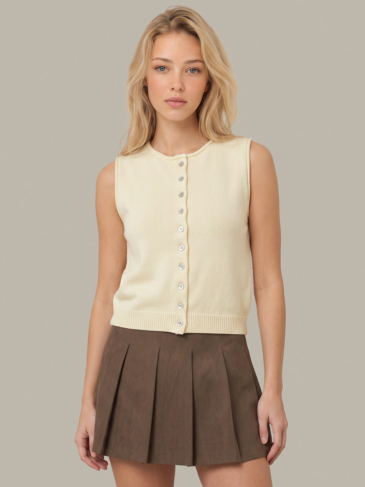A blonde model wearing a cream sleeveless button-front knit vest and a brown pleated mini skirt, standing against a solid light gray studio background.