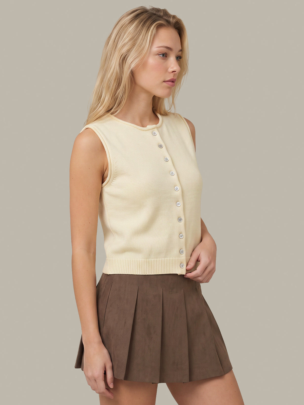 A blonde model wearing a cream sleeveless button-front knit top and a brown pleated mini skirt, posed against a solid light gray studio background.