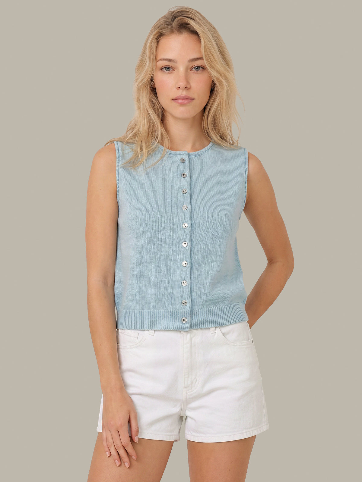 A woman modeling a light blue sleeveless knit button-front top with white shorts against a solid taupe background.