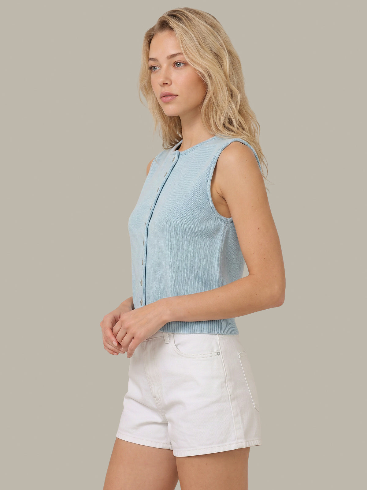 A woman wearing a light blue sleeveless knit vest with front buttons and white shorts, standing against a solid beige background.