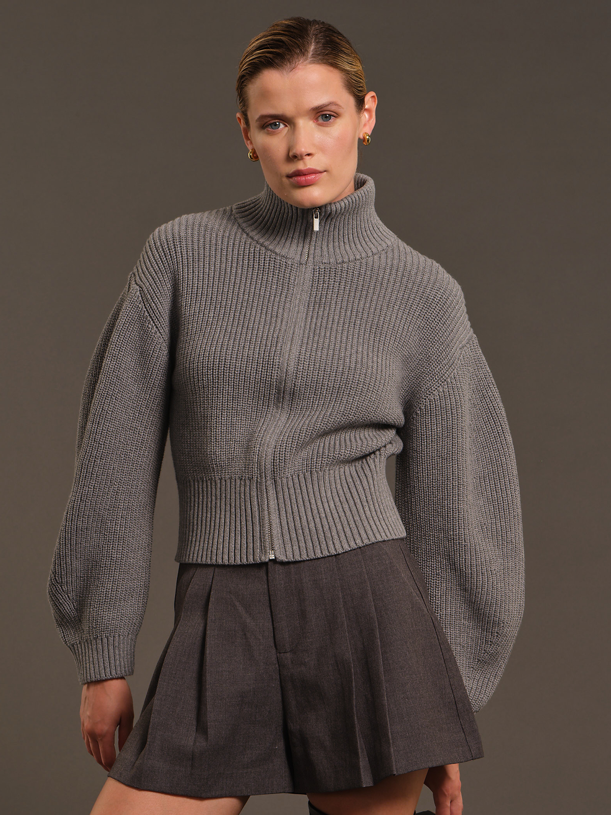 A model wearing a gray ribbed mock-neck zip-up sweater with balloon sleeves and a dark gray pleated skirt, set against a solid dark gray studio background.