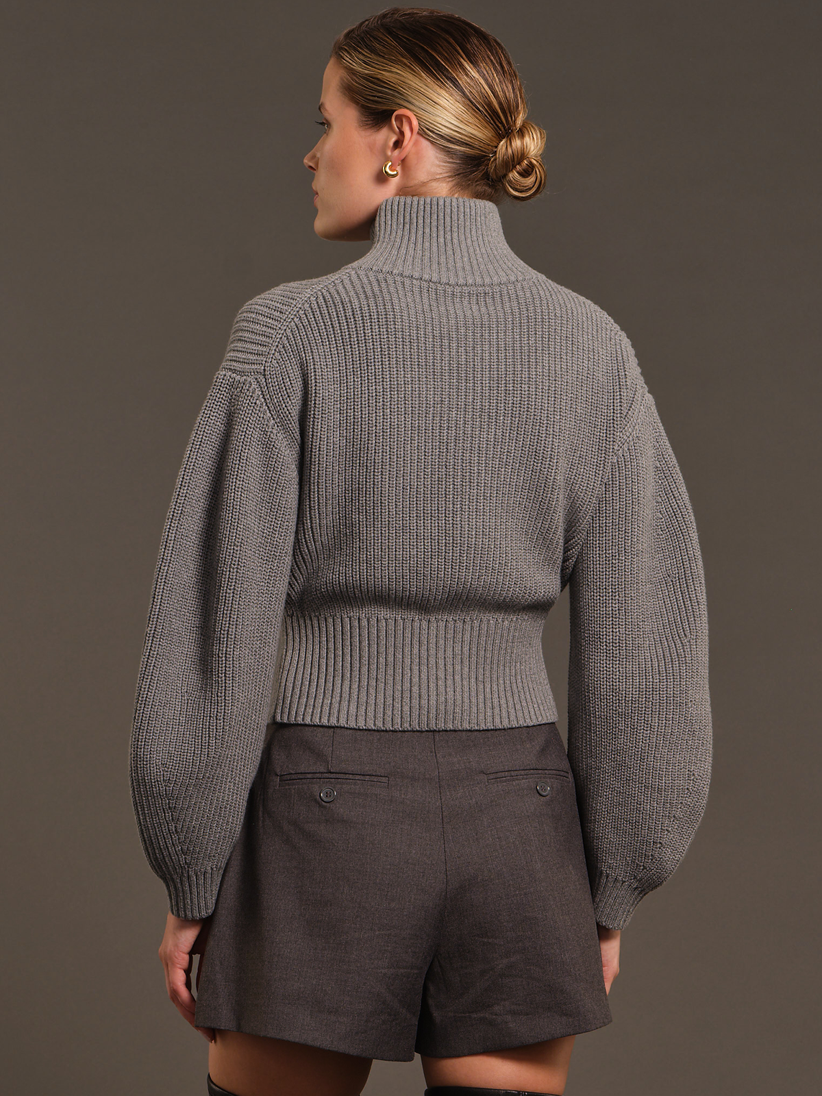 A woman shown from the back wearing a ribbed gray turtleneck sweater with voluminous sleeves and dark gray shorts, photographed against a plain taupe studio background.