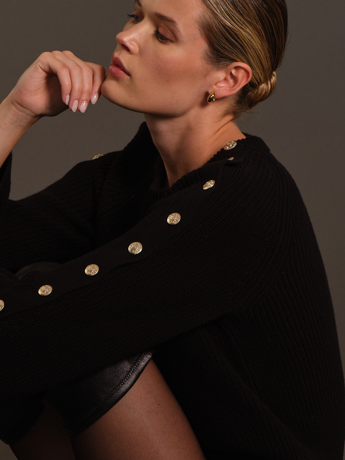 A close-up lifestyle shot of a model wearing a black ribbed knit sweater with prominent gold buttons along the collar and sleeve, set against a dark gray studio background.