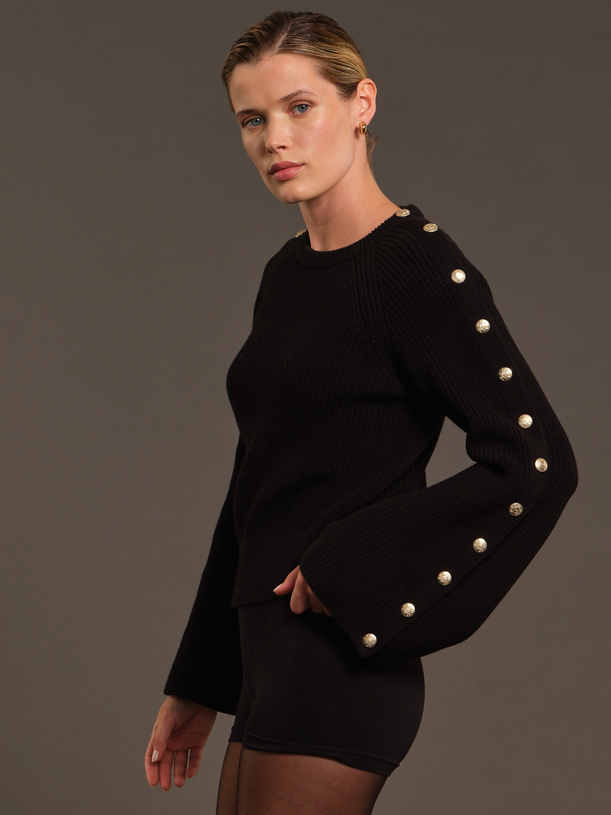 A model wearing a black ribbed knit sweater with decorative gold-toned buttons down the sleeves, paired with a matching black mini skirt, against a solid dark gray studio background.