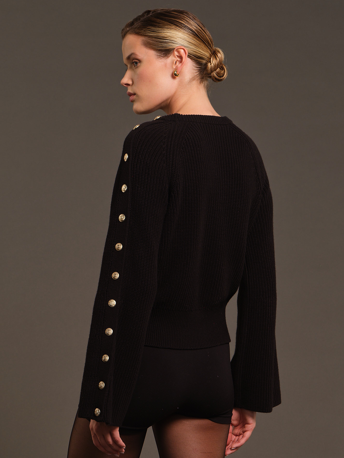 A woman shown from the back wearing a black ribbed knit sweater with a row of prominent gold buttons down the back, paired with black shorts, against a solid dark gray studio background.