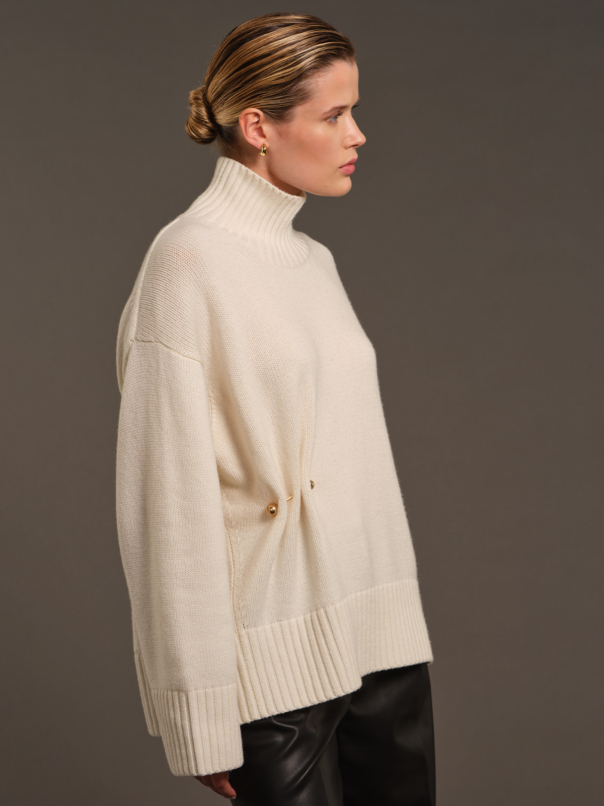 A woman in side profile wearing a cream-colored knit turtleneck sweater with a buttoned front detail and ribbed cuffs, paired with black leather pants, against a solid dark gray studio background.