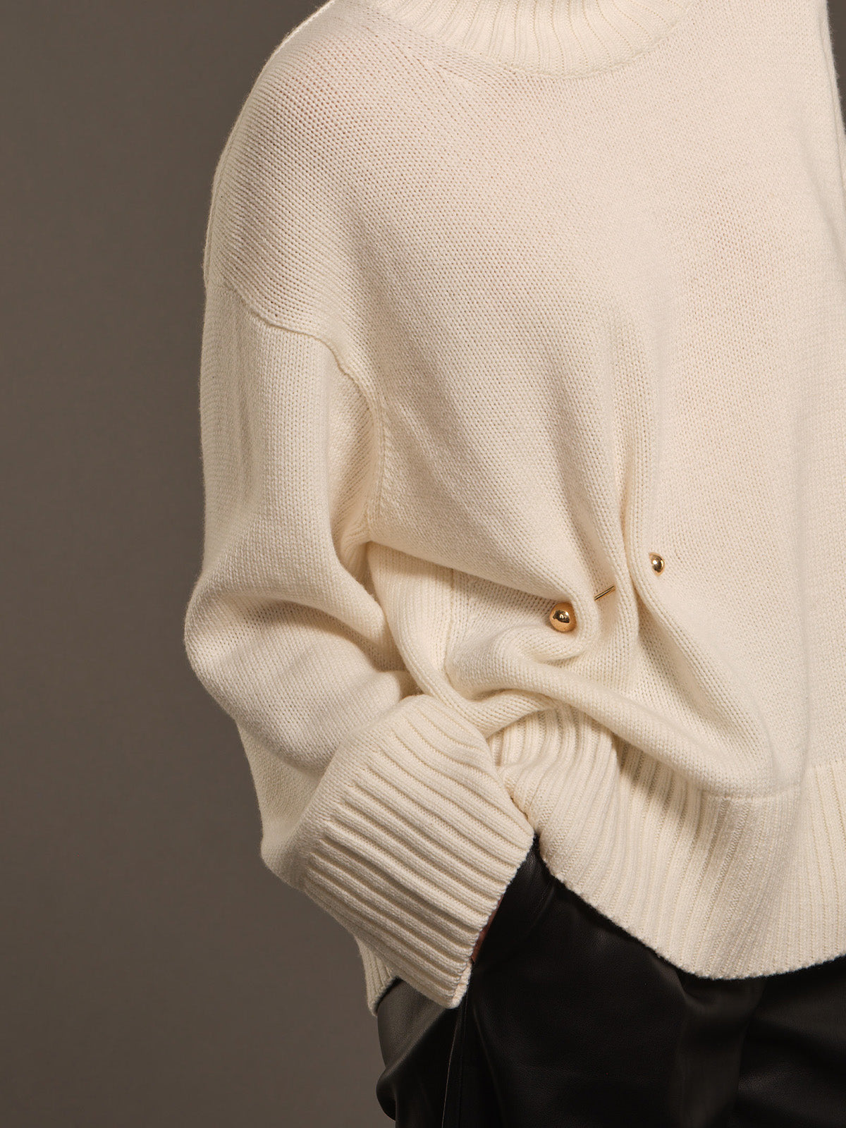 Close-up of a cream-colored knit sweater with a crew neck, ribbed cuffs, and decorative gold buttons, paired with black trousers against a dark gray background.