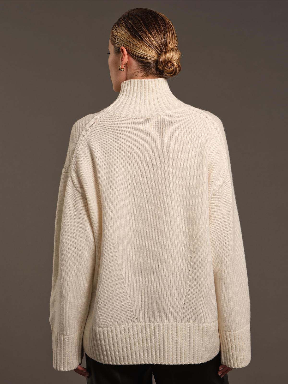 A woman viewed from the back wearing an oversized cream-colored knit turtleneck sweater, paired with black pants, against a solid dark gray studio background.