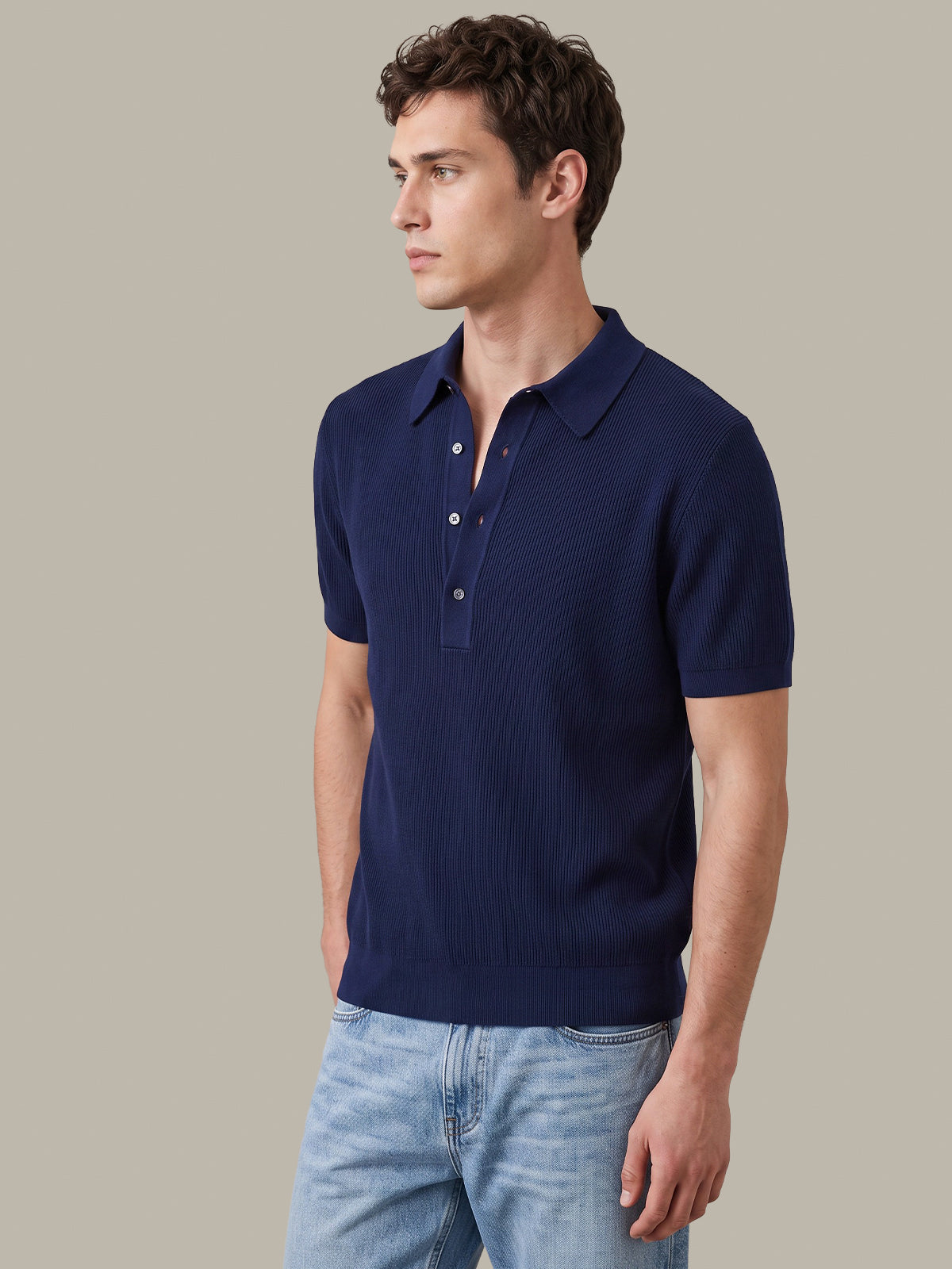 A man wearing a navy blue short-sleeve knit polo shirt with a three-button placket, paired with light blue jeans, photographed against a solid neutral beige background.