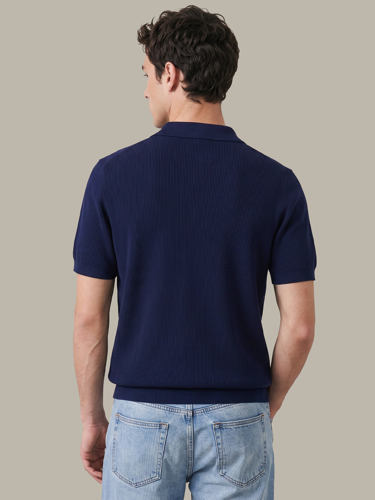 A man viewed from the back wearing a navy blue short-sleeve ribbed knit polo shirt and light blue jeans, set against a solid warm gray studio background.