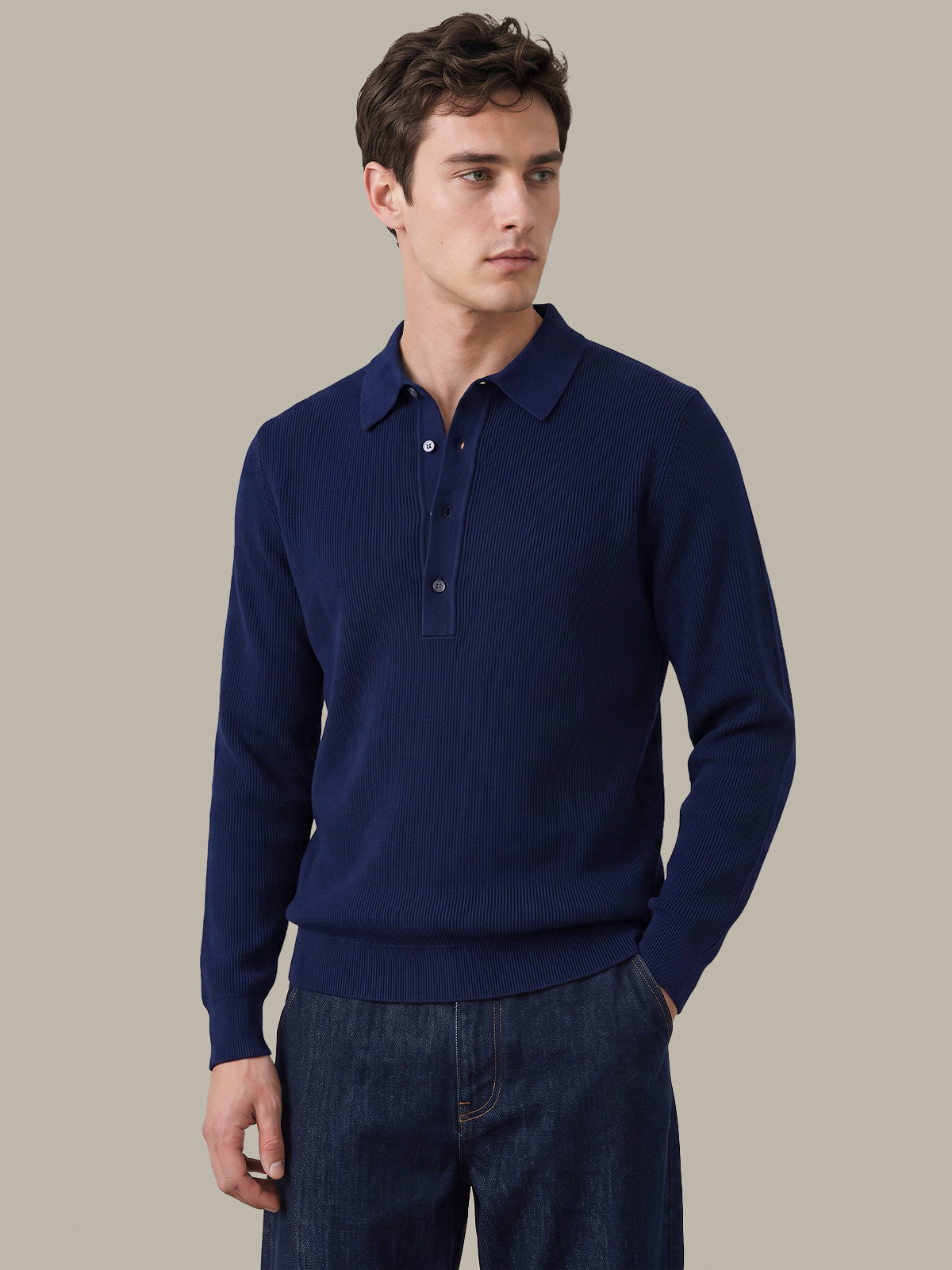 A man wearing a navy blue long-sleeve polo sweater and dark blue jeans, standing against a solid light gray background.
