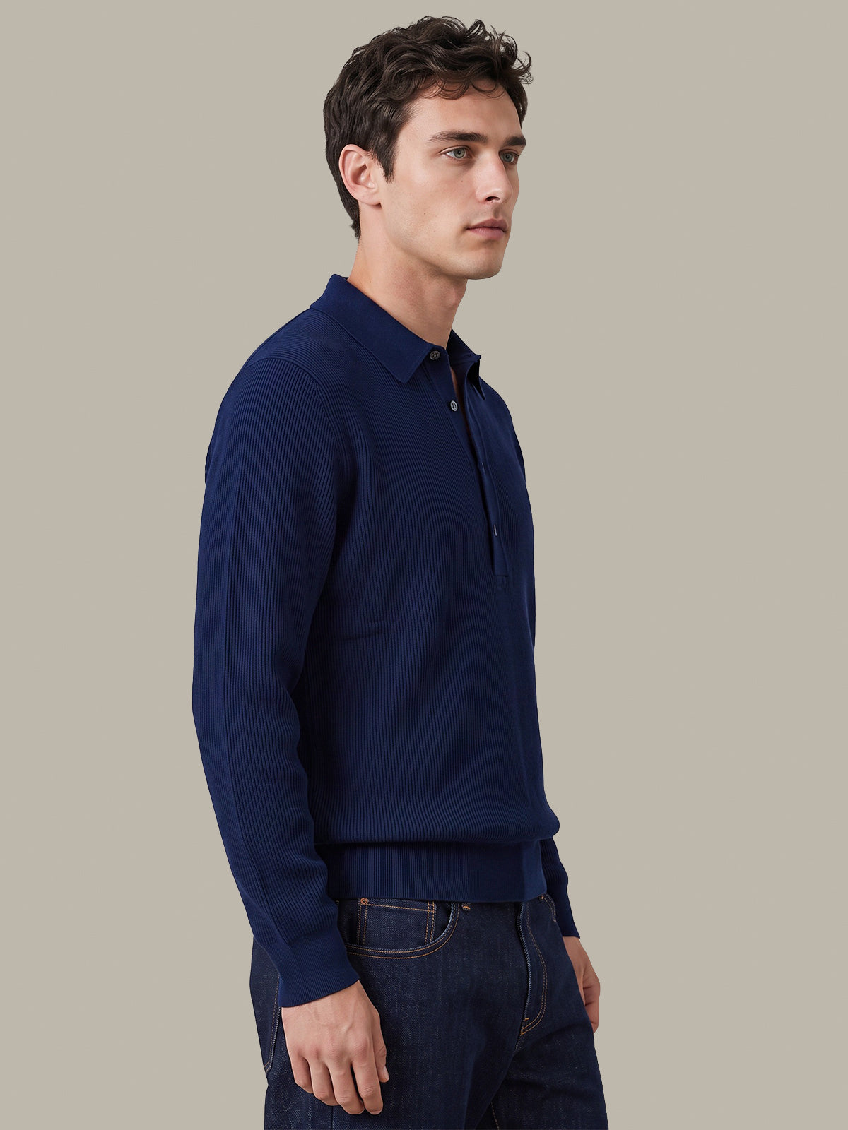 A man wearing a navy blue long-sleeve polo shirt and dark blue jeans, standing in profile against a solid light gray background.