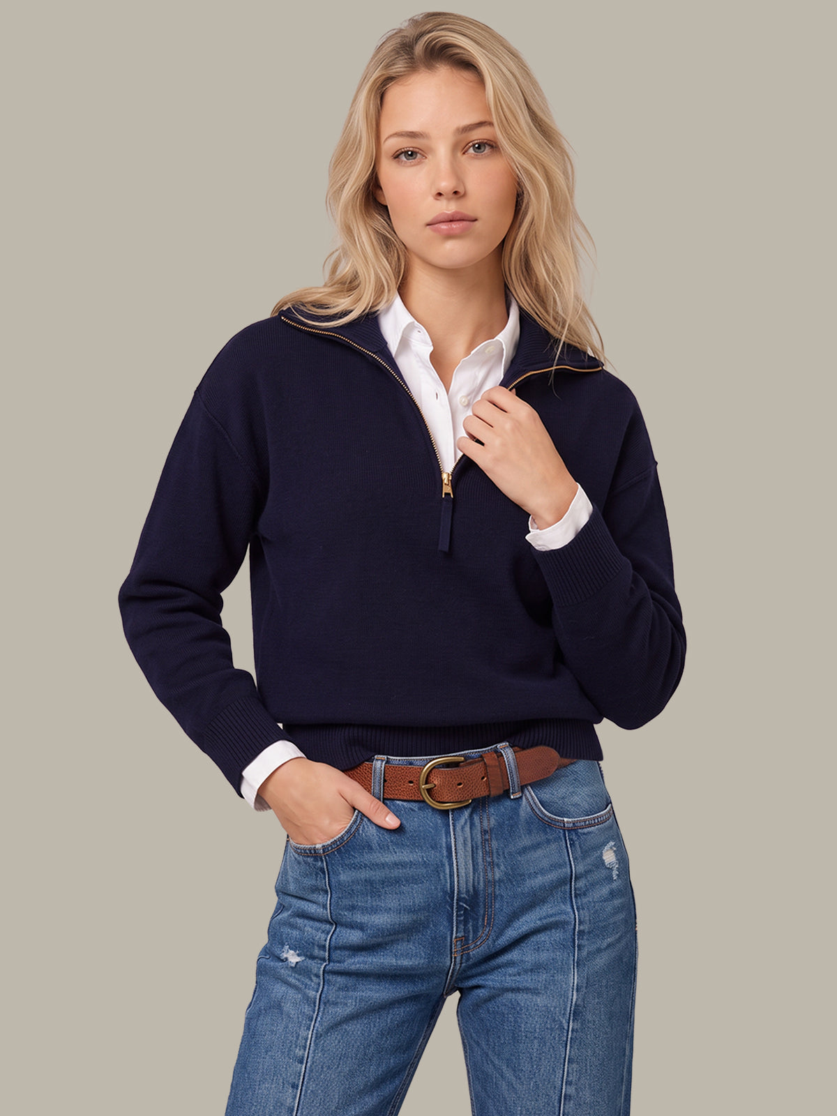 A woman modeling a navy blue quarter-zip sweater over a white collared shirt, paired with blue jeans and a brown belt, against a solid light gray background.