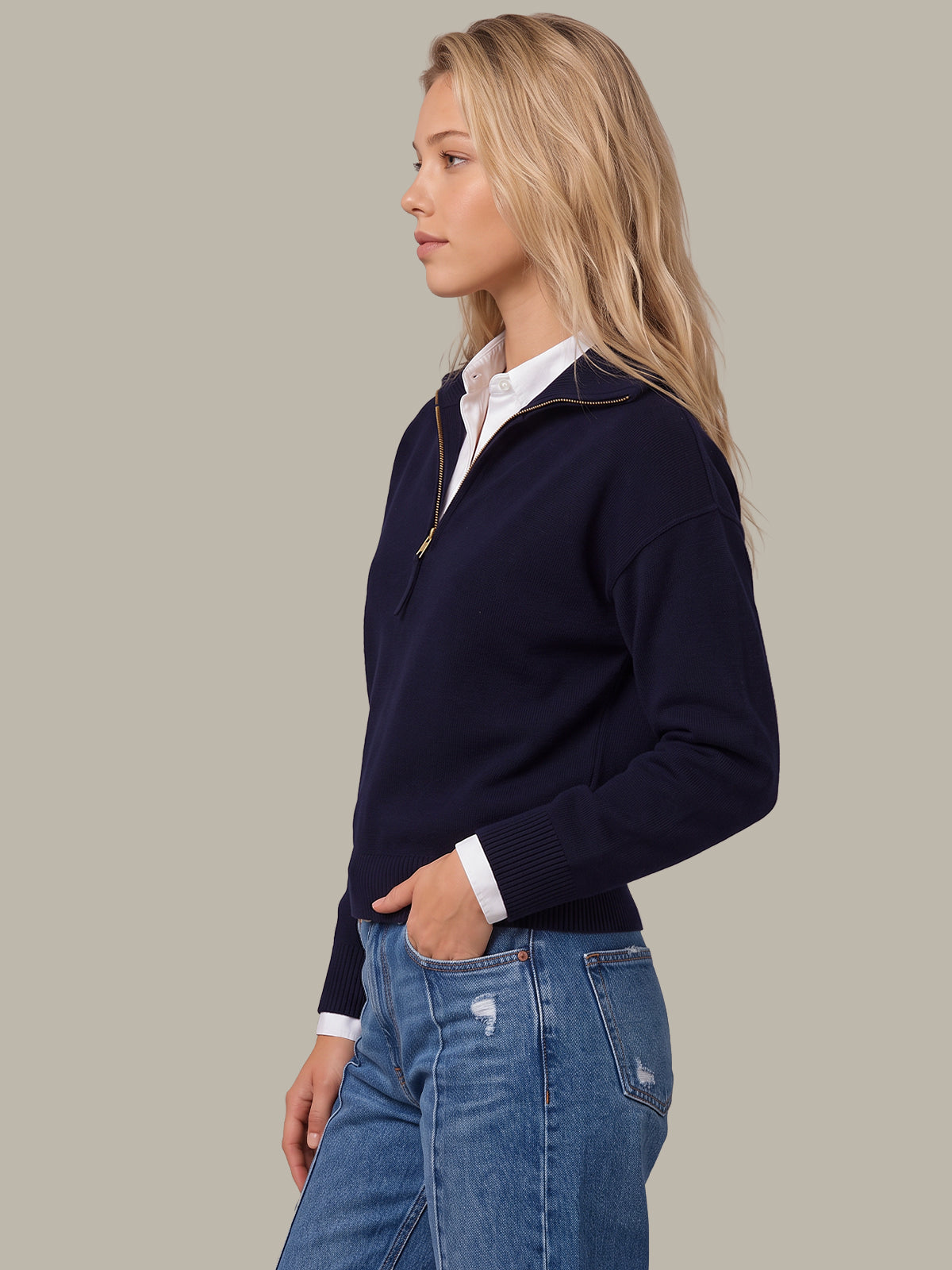 A woman in side profile wearing a navy blue quarter-zip sweater over a white collared shirt and blue jeans, set against a solid light gray background.