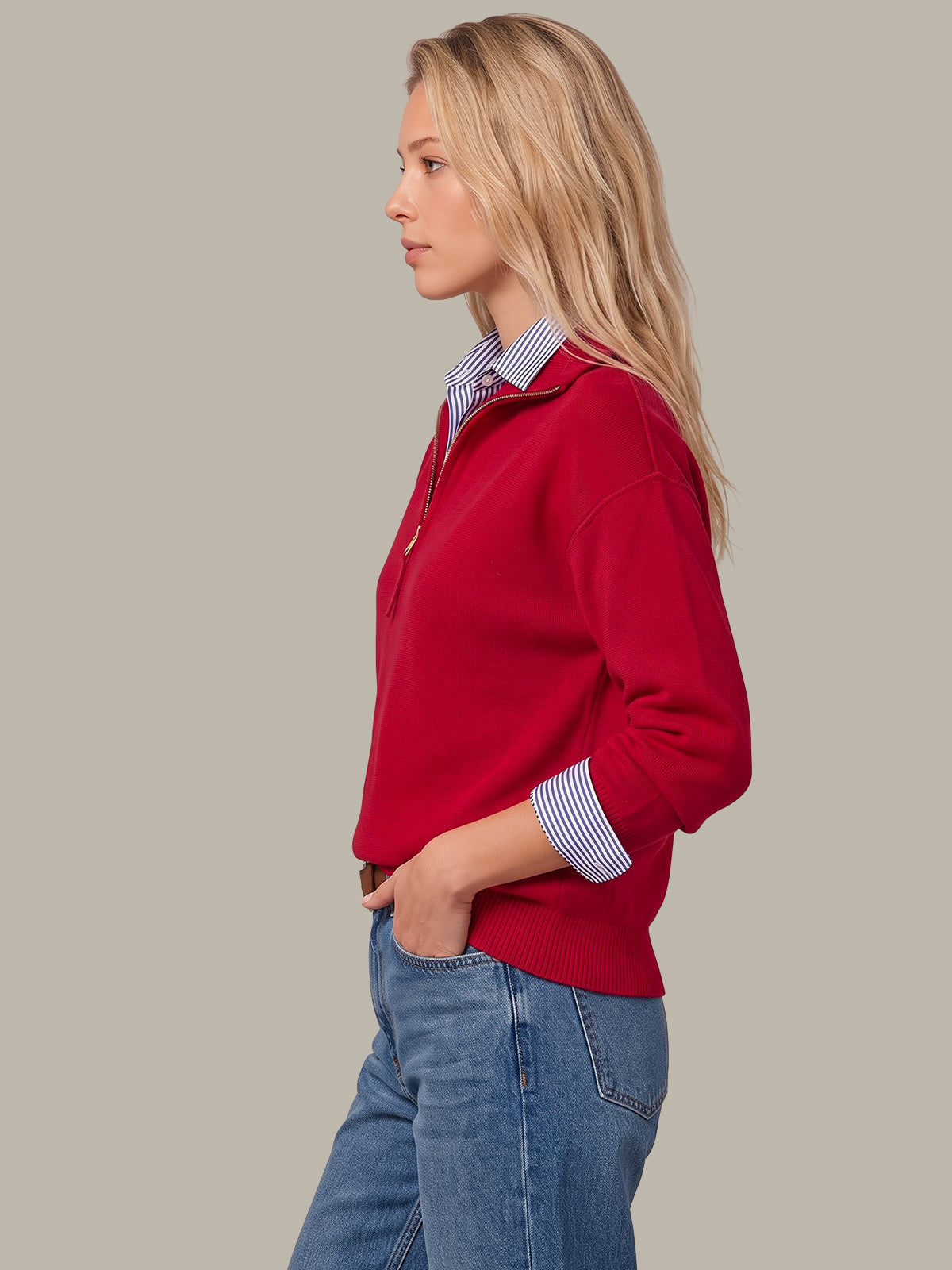 A woman in profile wearing a red quarter-zip knit sweater over a blue and white striped collared shirt, paired with blue jeans, against a solid light gray background.