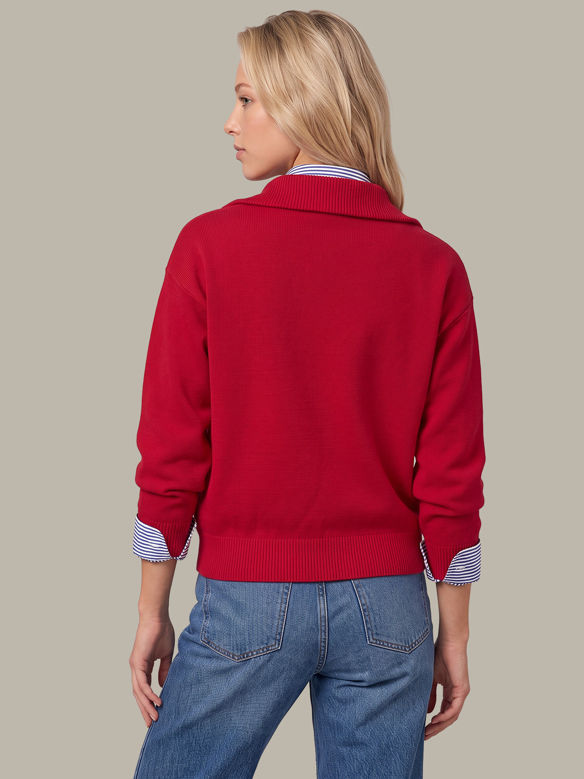 A woman shown from the back wearing a bright red knit sweater over a blue and white striped collared shirt, paired with blue jeans, against a solid taupe background.