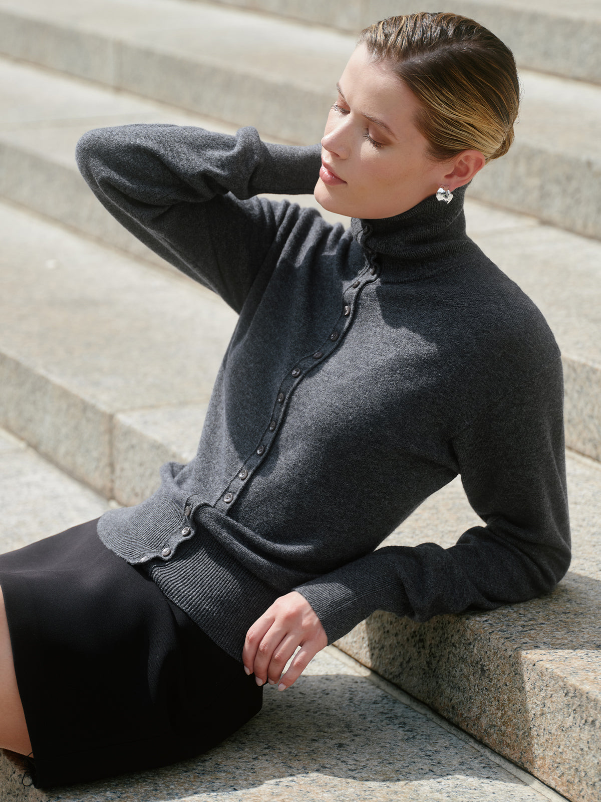 A woman wearing a dark gray ribbed turtleneck cardigan with scalloped edges and a button front, paired with a black skirt, sitting on stone steps.