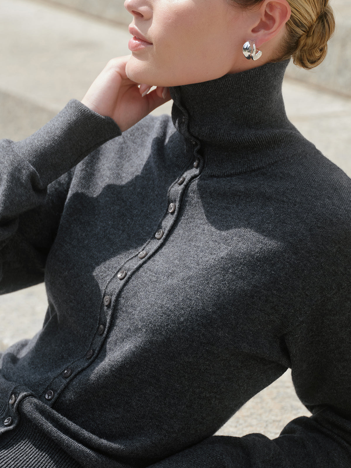 A close-up of a model wearing a dark gray turtleneck cardigan with a buttoned front placket, set against a blurred outdoor background.