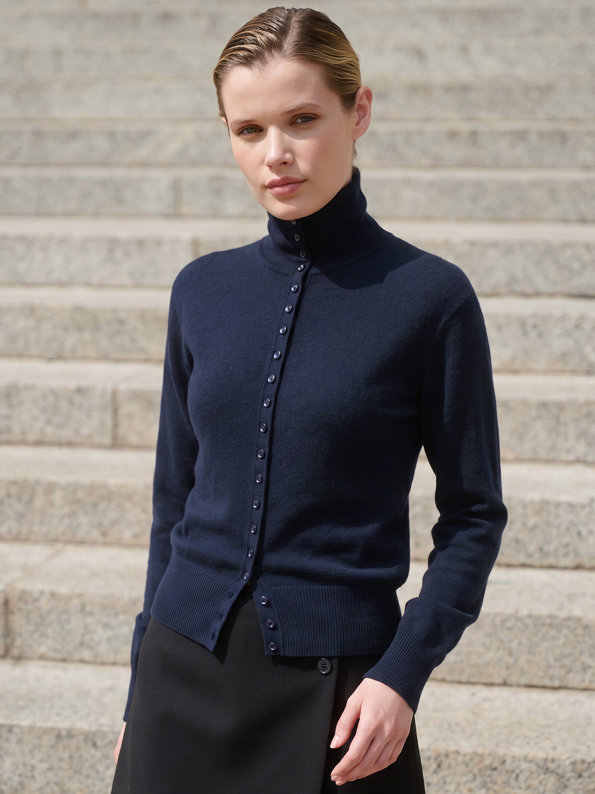 A woman wearing a navy blue mock-neck button-front cardigan sweater and a black skirt, standing in front of outdoor stone steps.