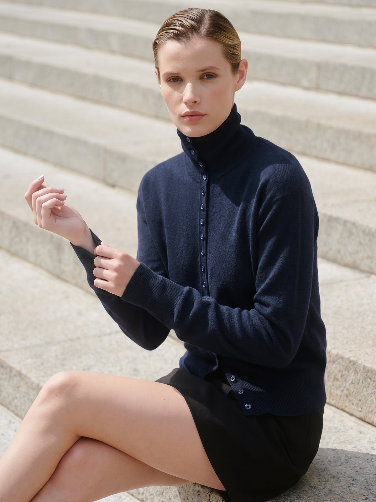 A woman sits on stone steps wearing a navy blue mock-neck button-front cardigan and a black skirt.