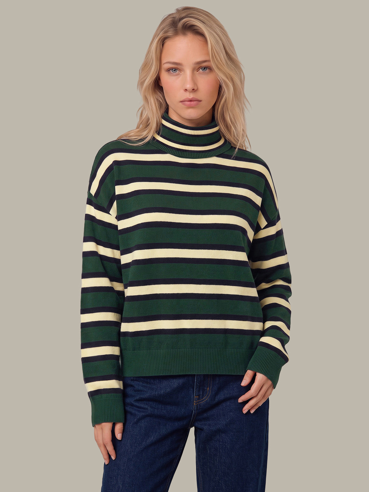 A woman wearing a dark green and cream striped turtleneck sweater with dark blue jeans, standing against a solid light gray background.