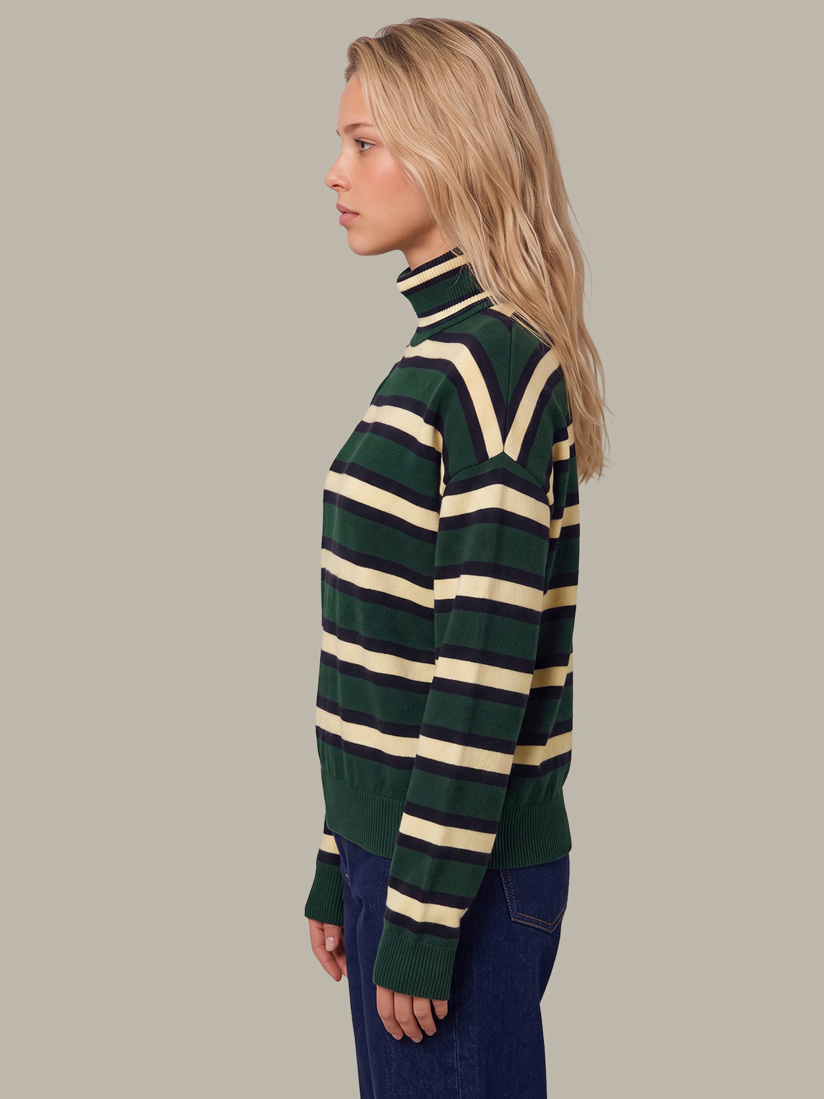 A woman shown in side profile wearing a green and cream striped turtleneck sweater and dark blue jeans, set against a solid light gray background.