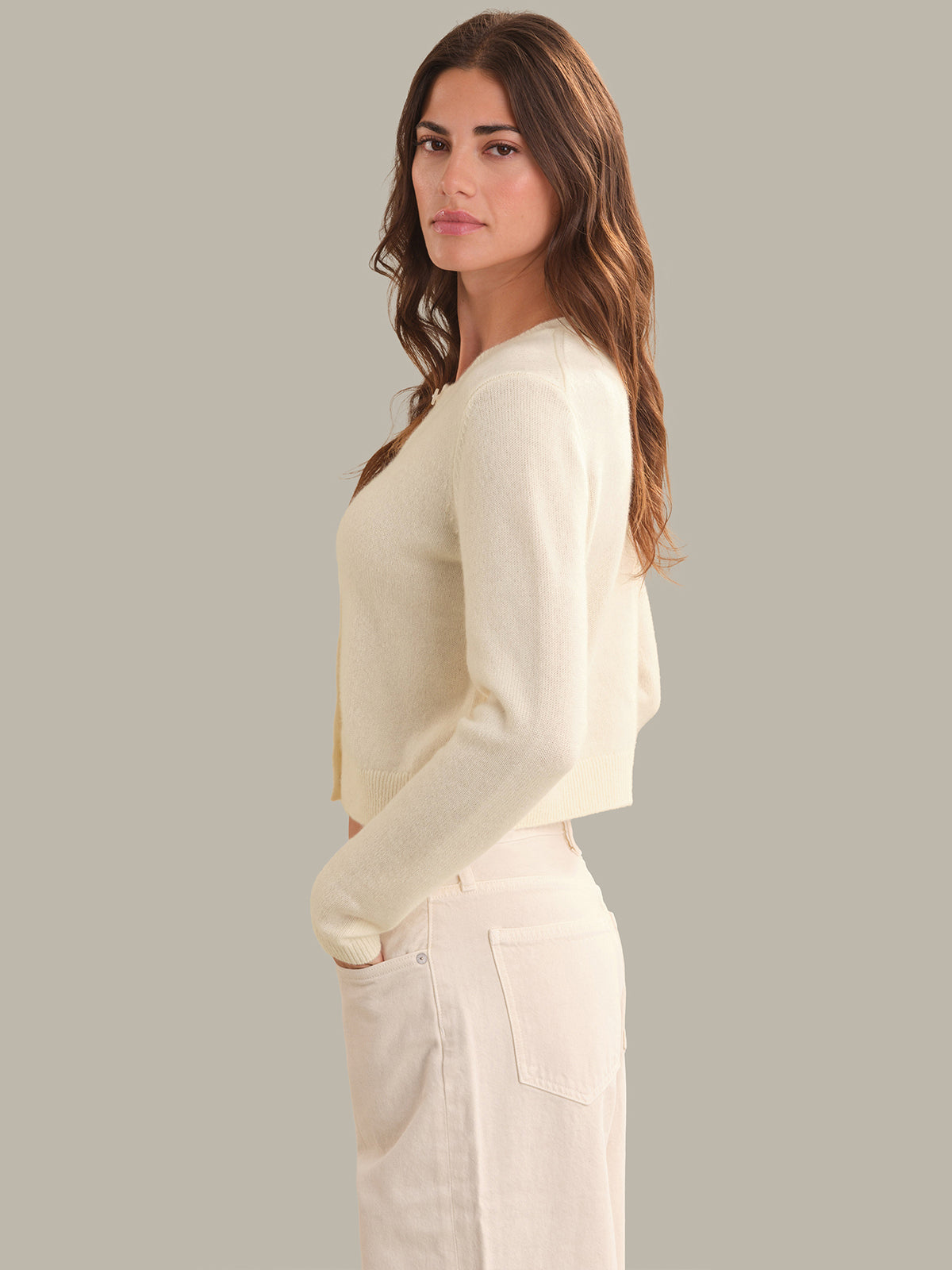 A woman modeling a cream-colored knit cardigan sweater paired with matching cream pants, shown from a side profile against a solid light gray background.