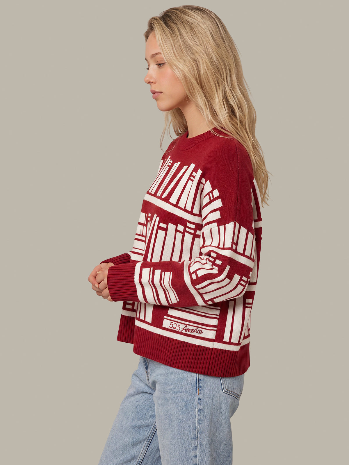A woman in profile wearing a red and white patterned knit sweater with light blue jeans, set against a solid gray studio background.