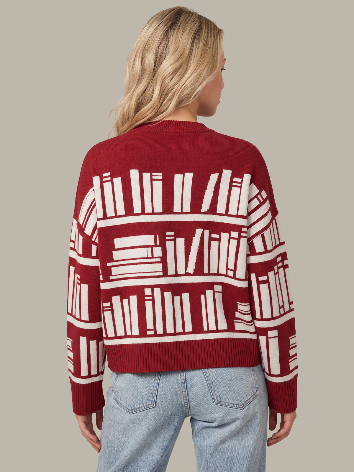 A woman viewed from the back wearing a red knit sweater with a white bookshelf pattern and light blue jeans, set against a solid gray background.