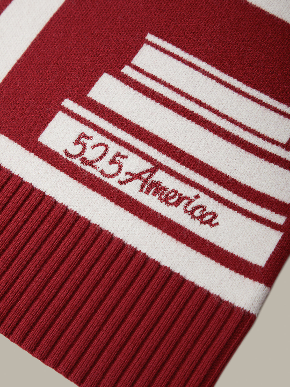 Close-up of a knit beanie in deep red and white, featuring horizontal stripes, a ribbed cuff, and the embroidered text '525 America'.