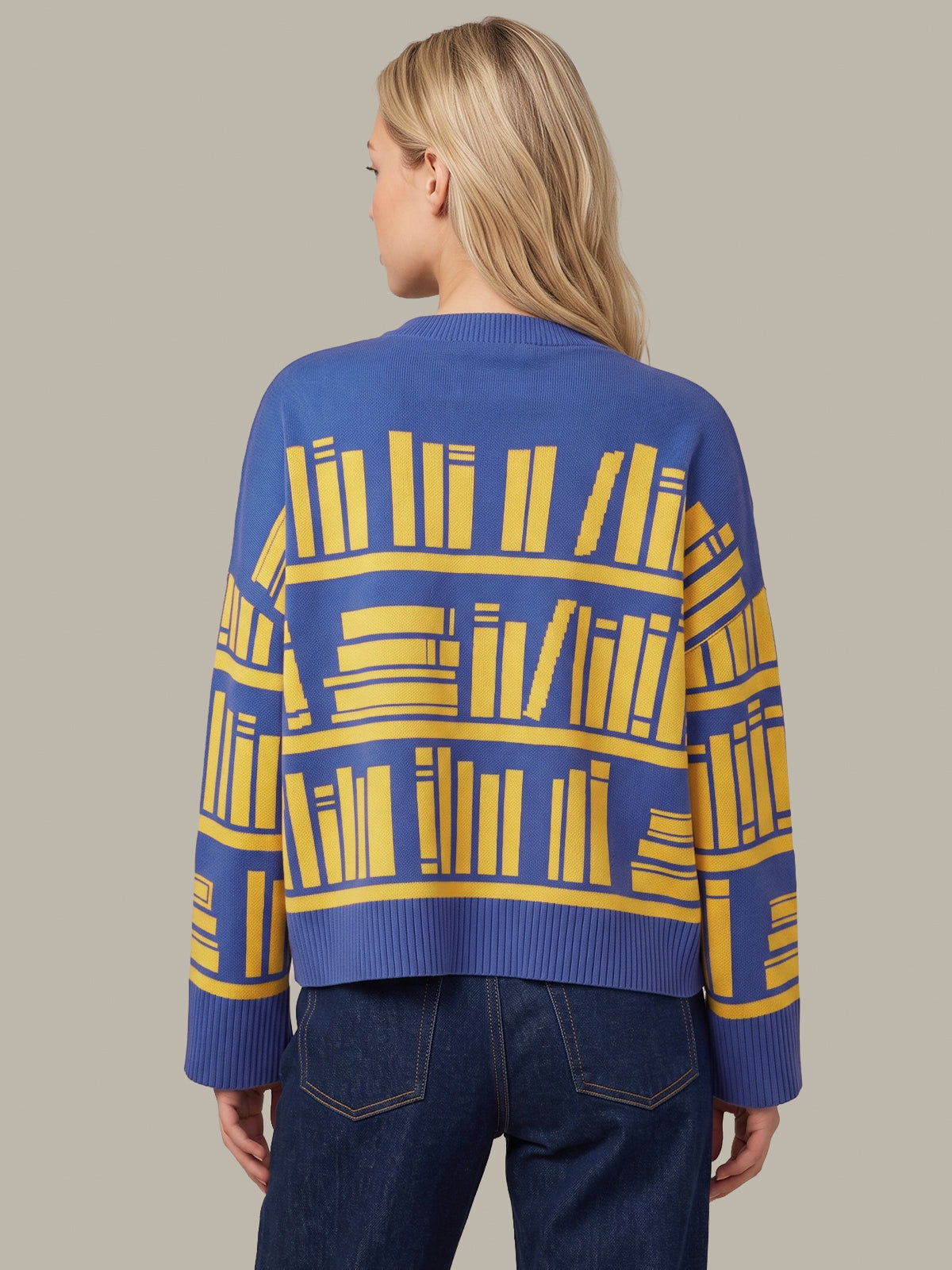 A woman viewed from the back wearing a blue knit sweater with a yellow bookshelf pattern, paired with dark blue jeans, against a solid light gray background.