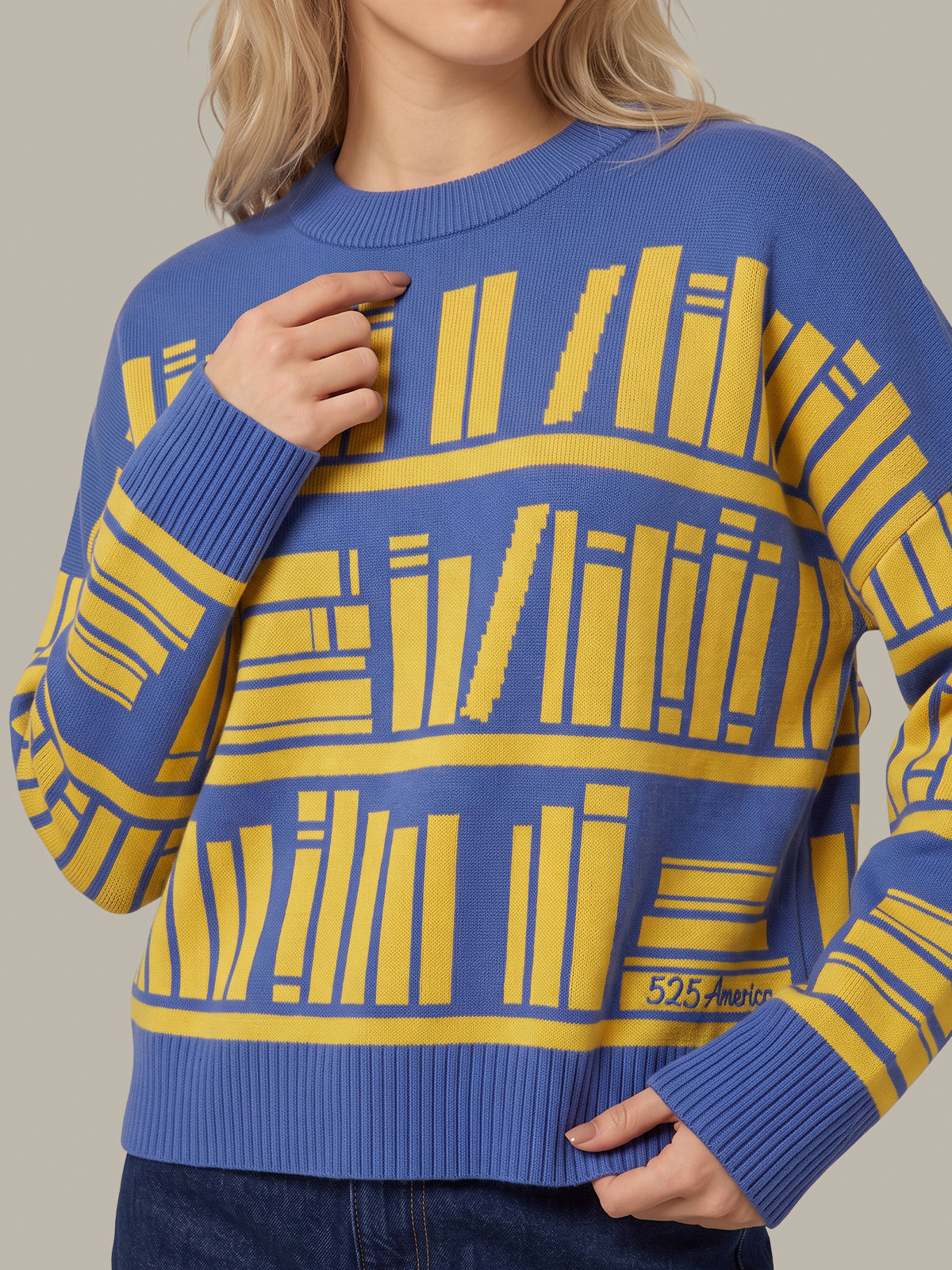 A close-up of a person wearing a blue and yellow knit sweater with a repeating bookshelf pattern, paired with dark blue jeans against a plain beige background.
