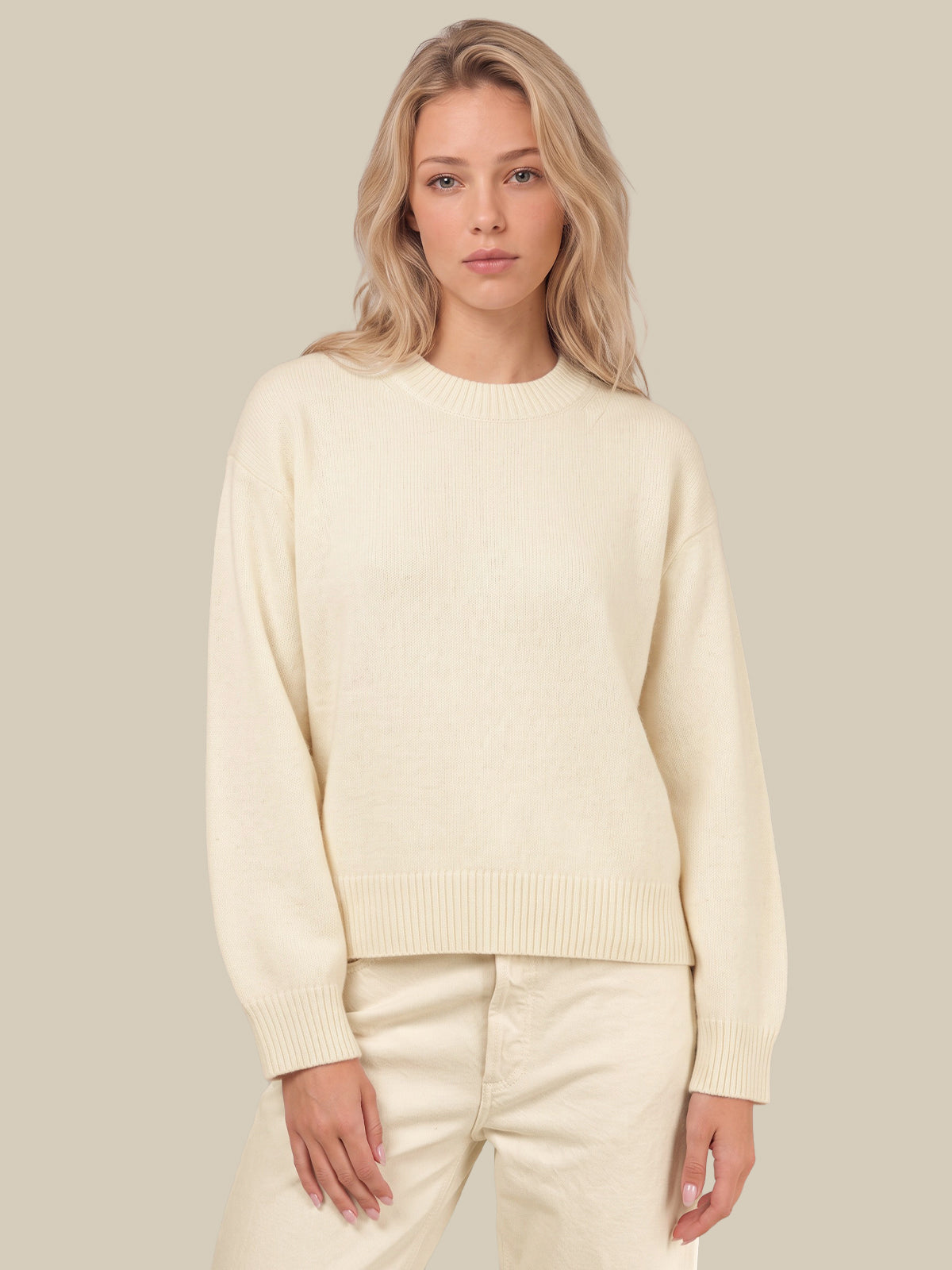 A woman wearing a cream-colored knit crewneck sweater and matching cream pants, standing against a solid light beige background.