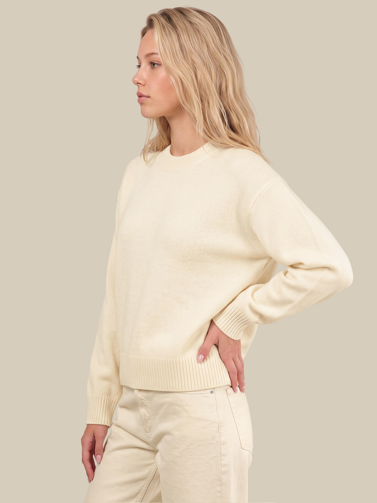A woman wearing a cream-colored crewneck sweater and matching cream pants, shown from a side profile against a solid light beige background.
