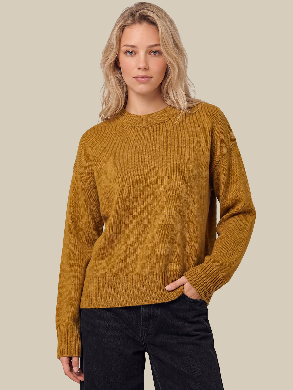 A woman wearing a mustard yellow ribbed knit sweater and dark jeans, standing against a solid light beige background.