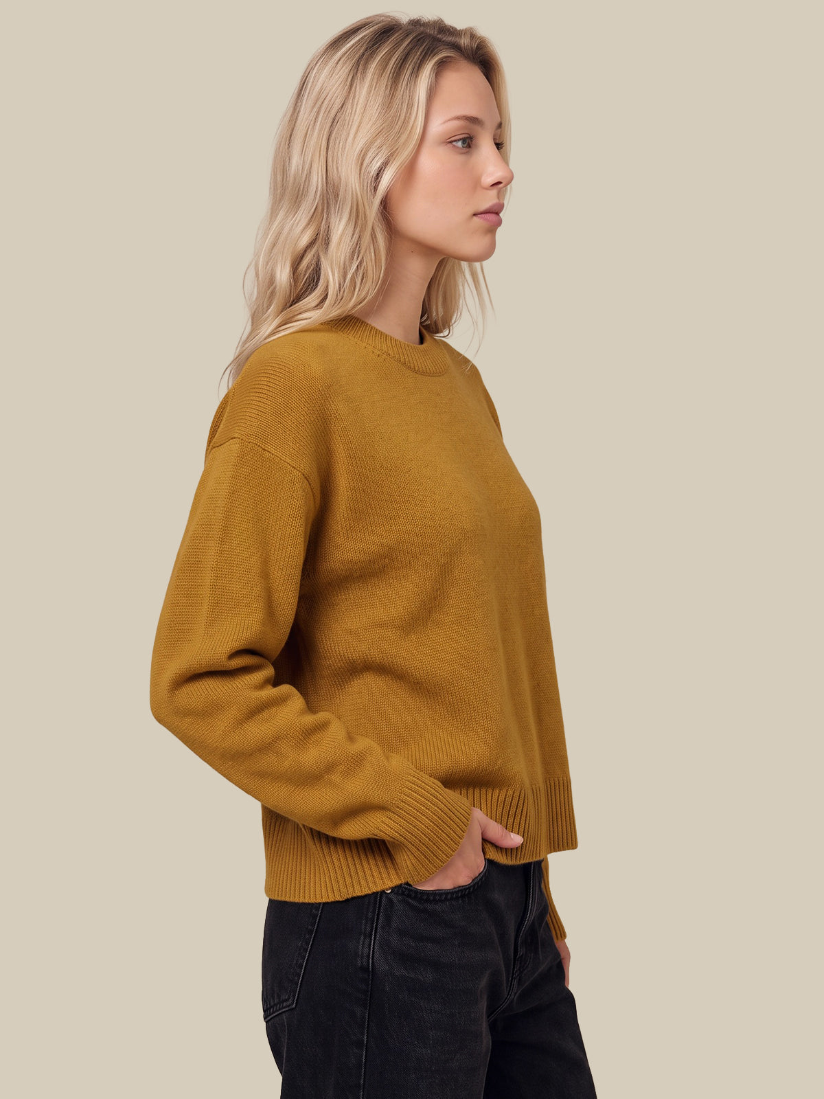 A woman in profile wearing a mustard yellow ribbed knit sweater and dark jeans, set against a solid light beige background.