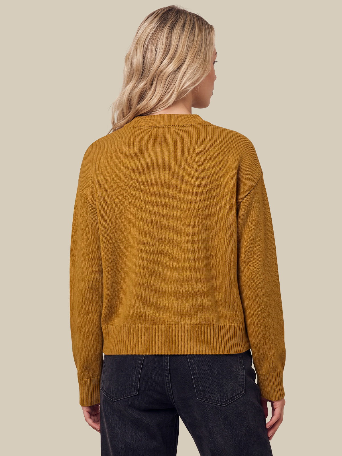 A woman viewed from the back wearing a mustard yellow knit sweater and dark jeans, set against a solid beige background.