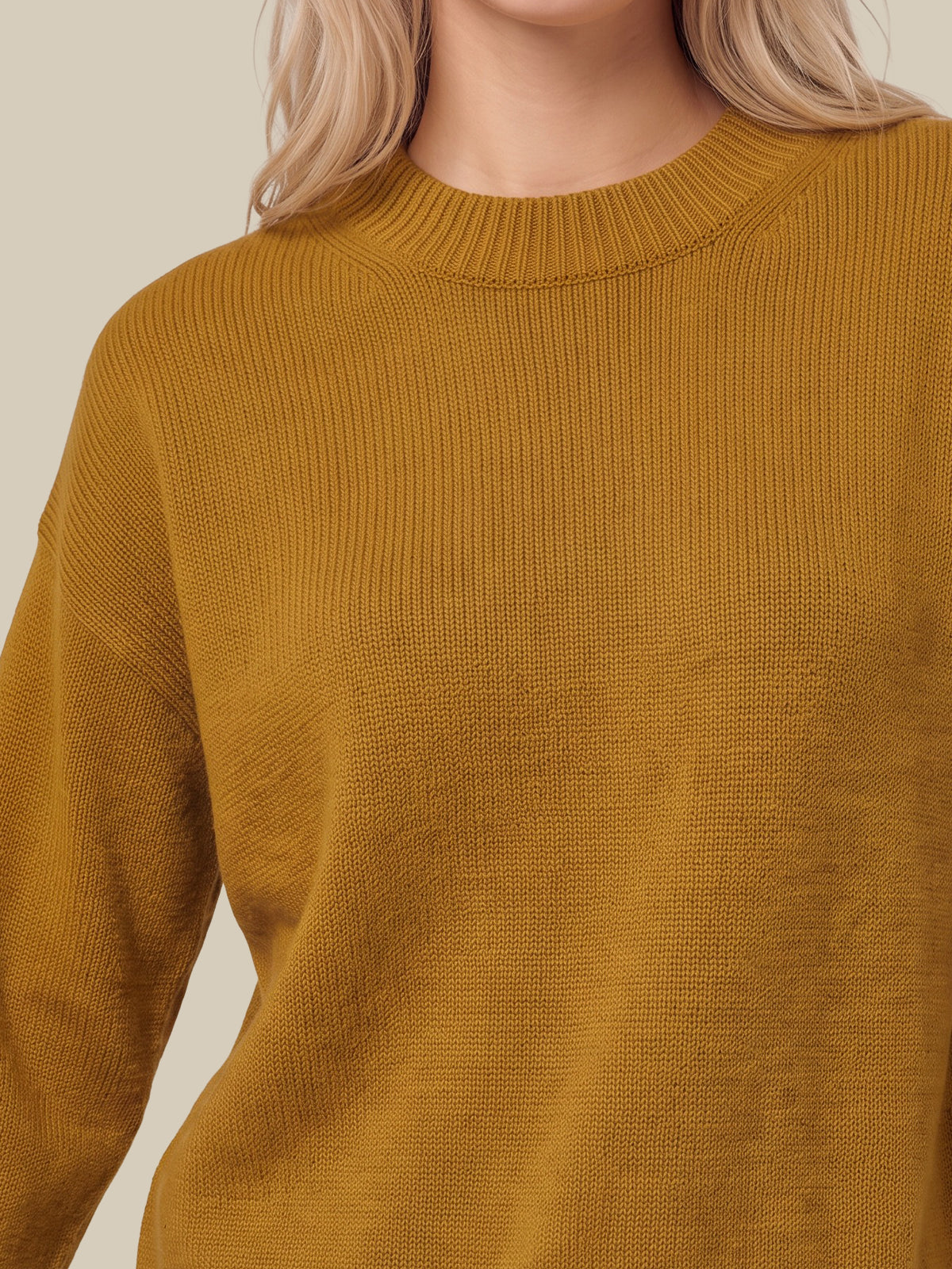 A close-up of a person wearing a ribbed knit crewneck sweater in a warm mustard yellow color, set against a solid light beige background.