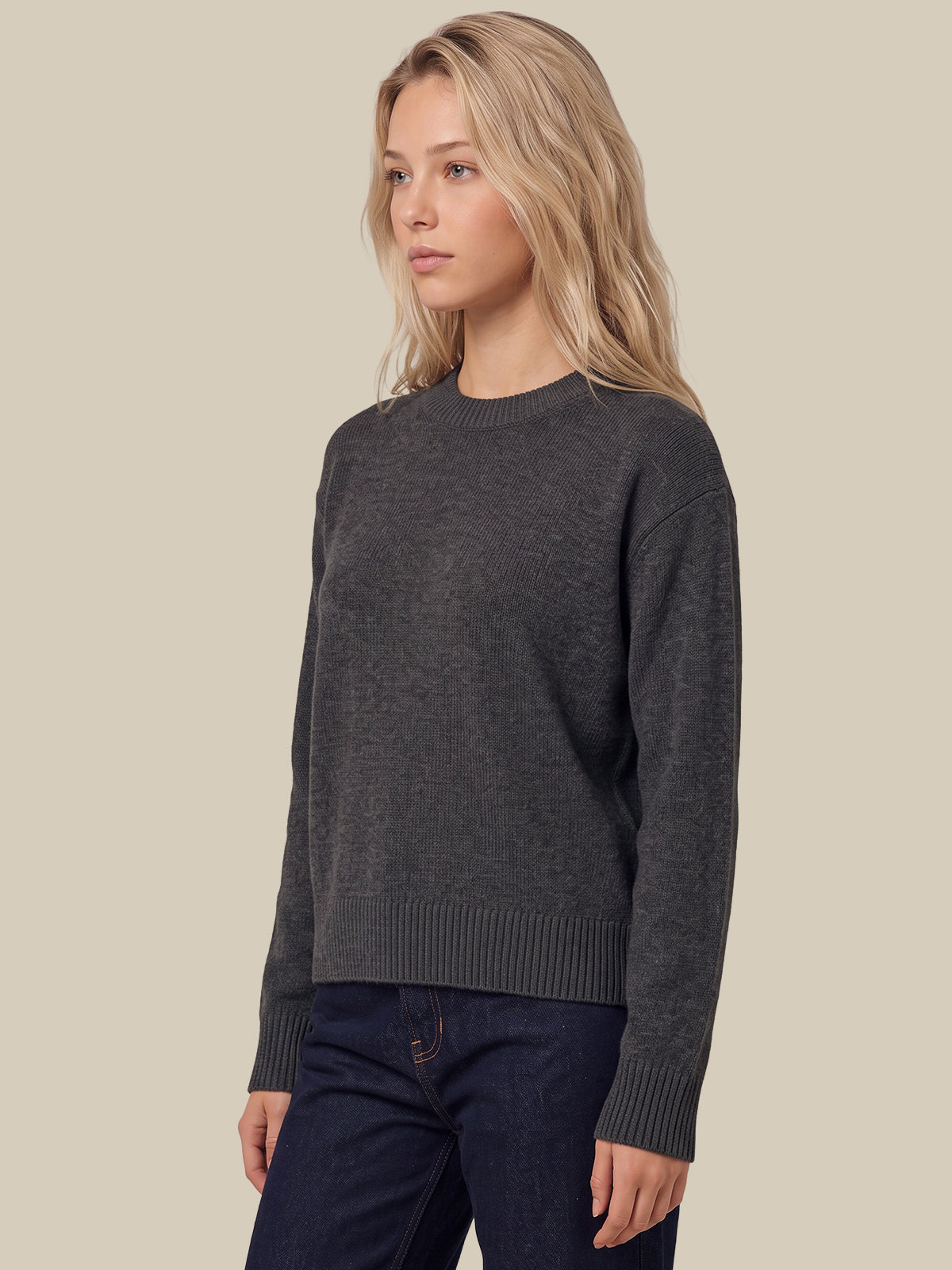 A woman wearing a dark charcoal gray knit crewneck sweater and dark blue jeans, standing against a solid beige background.