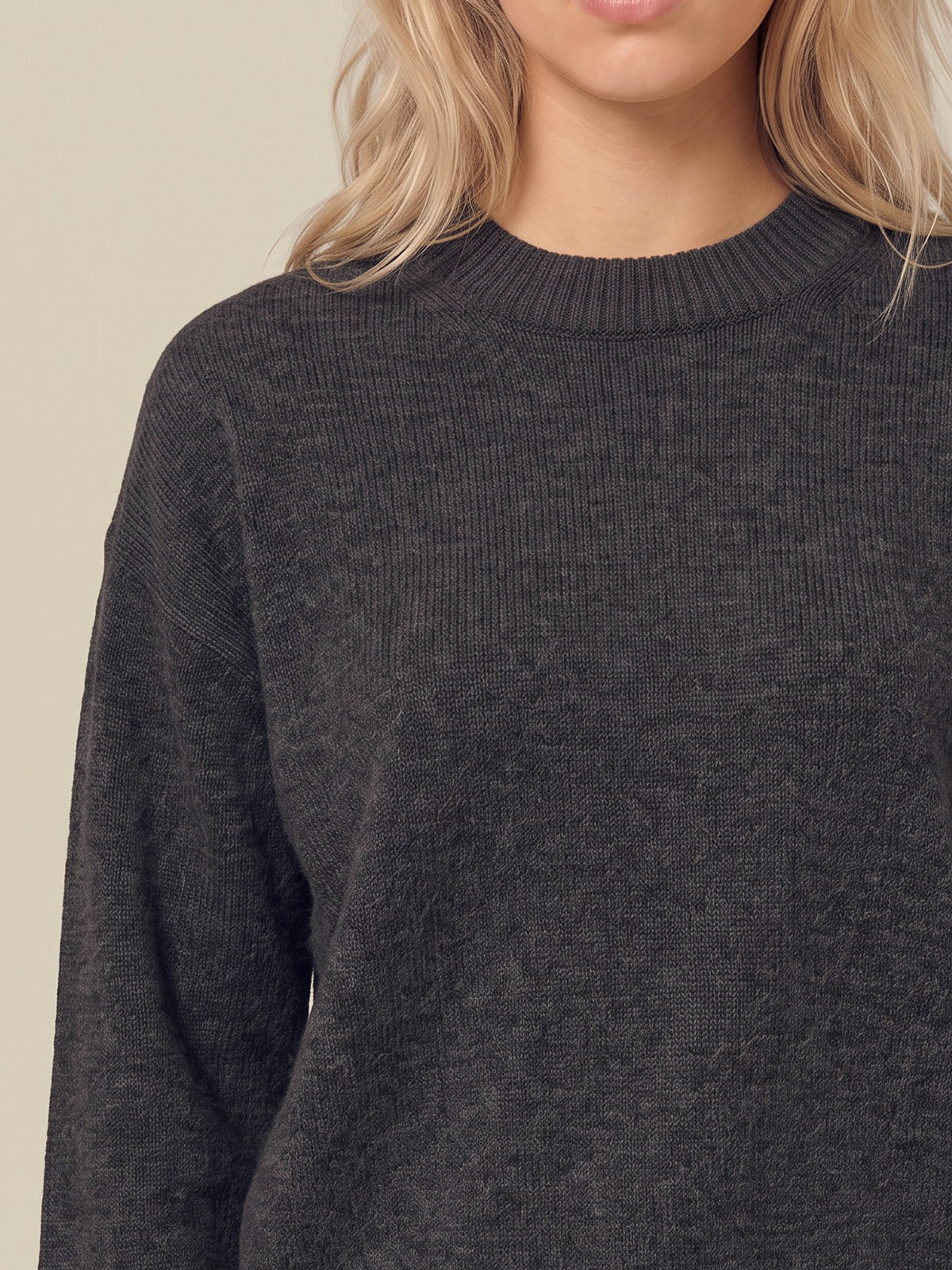 A close-up of a person wearing a dark charcoal gray ribbed knit crewneck sweater against a light beige background.
