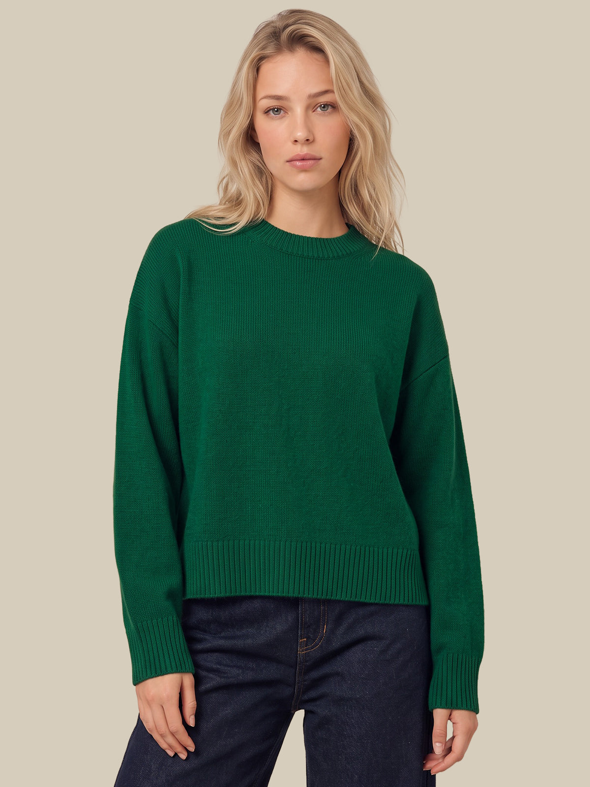 A blonde woman wearing a dark green crewneck knit sweater and dark blue jeans, standing against a solid beige background.