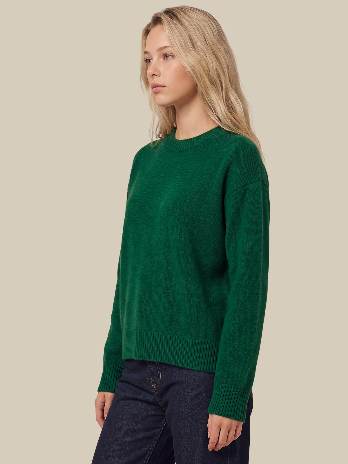 A woman wearing a dark green crewneck knit sweater and dark blue jeans, standing against a solid beige background.