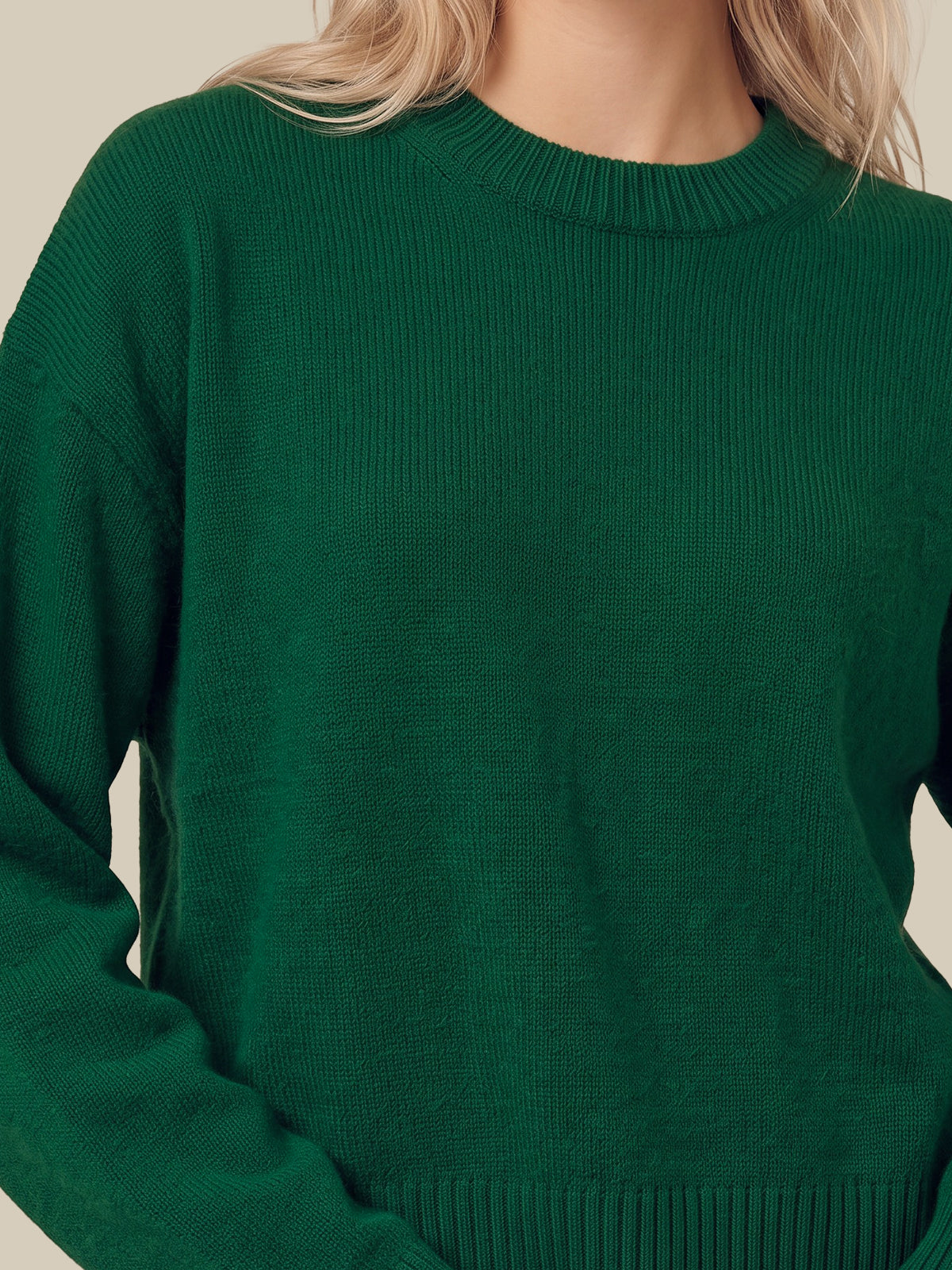 A close-up of a person wearing a dark green ribbed knit crewneck sweater, showing the texture and fit against a plain light beige background.