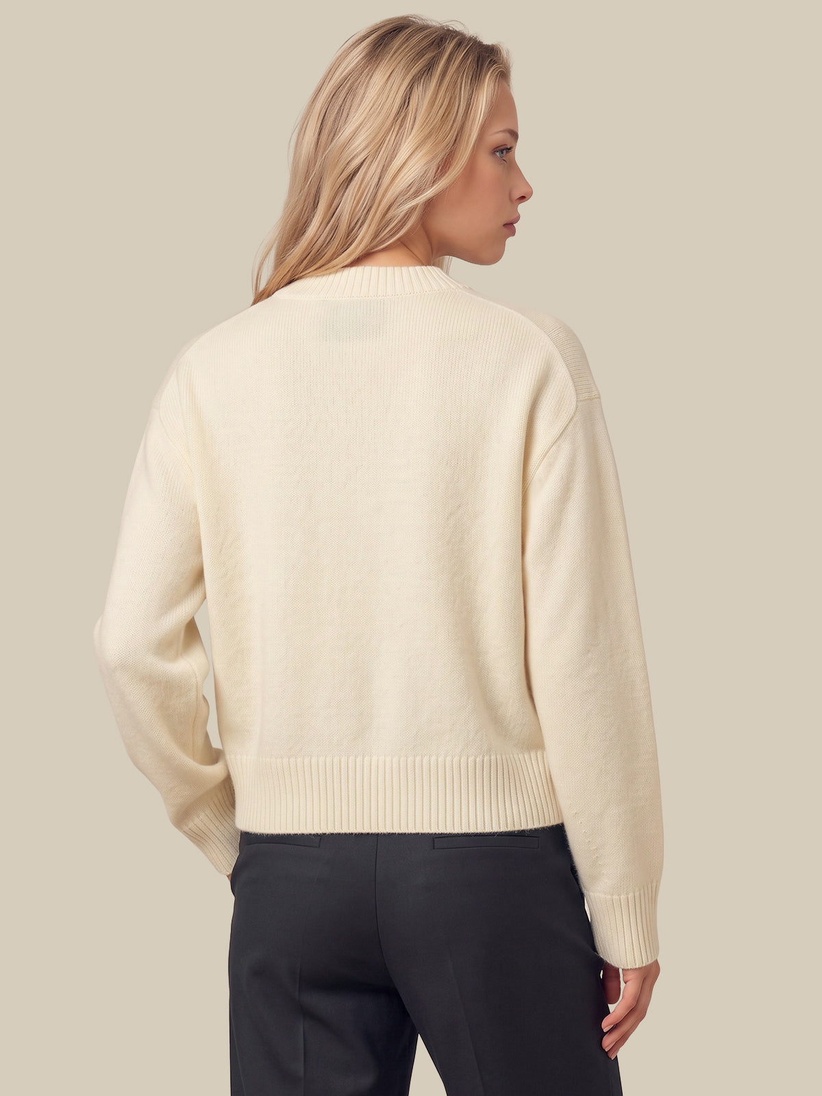 A woman shown from the back wearing a white-colored knit sweater and dark gray trousers, set against a solid light beige background.